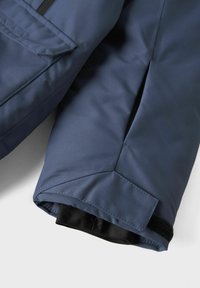 Marineblå ærme på jakken med en glat, tekstureret overflade. Har en Velcro-lukning ved cuffen og en vinkelret skæring for et moderne design.