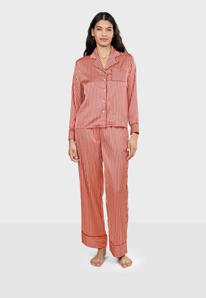 Femme debout pieds nus portant un haut de pyjama à manches longues boutonné, à rayures verticales rouges et blanches, et un pantalon ample assorti.