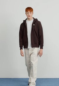 Sudadera marrón con cremallera y bolsillos, con un logo blanco, llevada sobre una camiseta blanca, combinada con pantalones beige claros, de pie sobre un suelo azul.