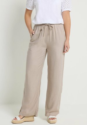 Vrouw draagt beige wijde broek met trekkoord, wit ooglet korte mouw shirt en witte sleehakken met enkelriempjes.