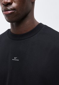 Κοντινό πλάνο ενός ατόμου που φοράει μαύρο μπλουζάκι Nike με το κείμενο «NIKE STANDARD ISSUE» και λογότυπο στο στήθος, σε ένα απλό φόντο.