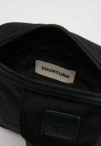 Indvendig af en sort stofpose med lynlåslukning. Har et mærke med teksten "YOURTURN" og et lille lædermærke. Enkel og funktionel design.