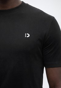 Μαύρο κοντομάνικο μπλουζάκι που φοριέται από άτομο, με μικρό λευκό αριστοτεχνικό λογότυπο "DC" στο πάνω αριστερό μέρος του στήθους.