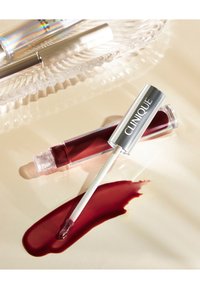 Clinique leppeprodukt med en gjennomsiktig tube; sølvhette, hvit applikator. Dyp rød farge på en lys overflate, blank tekstur.