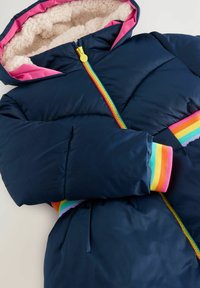 Tumši zilā pūkainā jaka ar rozā kapuci iekšpusi, varavīksnes krāsā svītrotiem aproču galiem un ar rāvējslēdzēju, kuram ir daudzkrāsains rāvējslēdzējs.