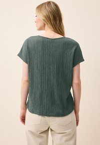 Top corto verde con texture e maniche corte, dal collo ampio e una vestibilità rilassata, visto da dietro, abbinato a pantaloni chiari.