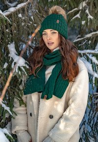 Berretto lavorato a maglia verde con un pom-pom di pelliccia e sciarpa abbinata, indossato con un cappotto in fleece beige, su uno sfondo di rami di sempreverdi imbiancati dalla neve.