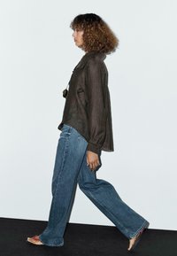 Chemise en lin marron à manches longues, coupe ample, associée à un jean flare en denim bleu taille haute. Des sandales à motif noir et rose soulignent le look décontracté.