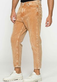 Pantaloni in velluto a coste color marrone chiaro con texture a righe verticali, vestibilità affusolata, cinque tasche e zip, abbinati a sneakers bianche.