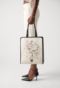 Canvas totebag med svarta handtag, prydd med modeskisser i röd och svart bläck. Rektangulär form, krämfärgad bakgrund och kontrasterande kanter.