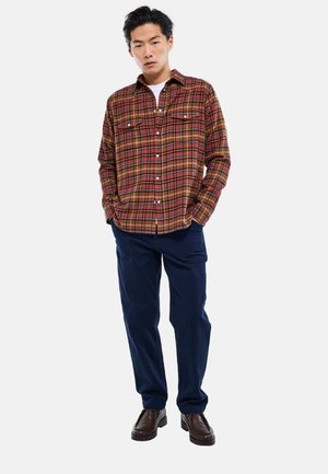 Jeune homme portant une chemise à carreaux marron et rouge boutonnée, un pantalon bleu marine et des chaussures marron foncé, debout les mains dans les poches.