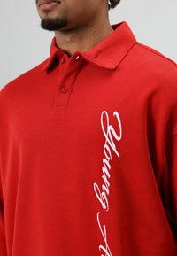 Rode polo met kraag, voorzien van twee knopen en witte geborduurde tekst met de woorden "Young." De stof heeft een gladde textuur.