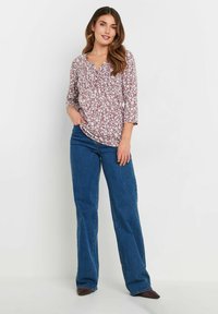 Bluse mit floralem Muster in Burgunderrot, mit V-Ausschnitt und Dreiviertelärmeln, kombiniert mit hochgeschnittenen blauen Schlagjeans und braunen Schuhen.