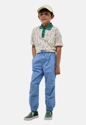 Polo à manches courtes avec col vert et motif de raquette de tennis, associé à un pantalon bleu à taille élastique et des chaussures vertes à enfiler. Casquette jaune.