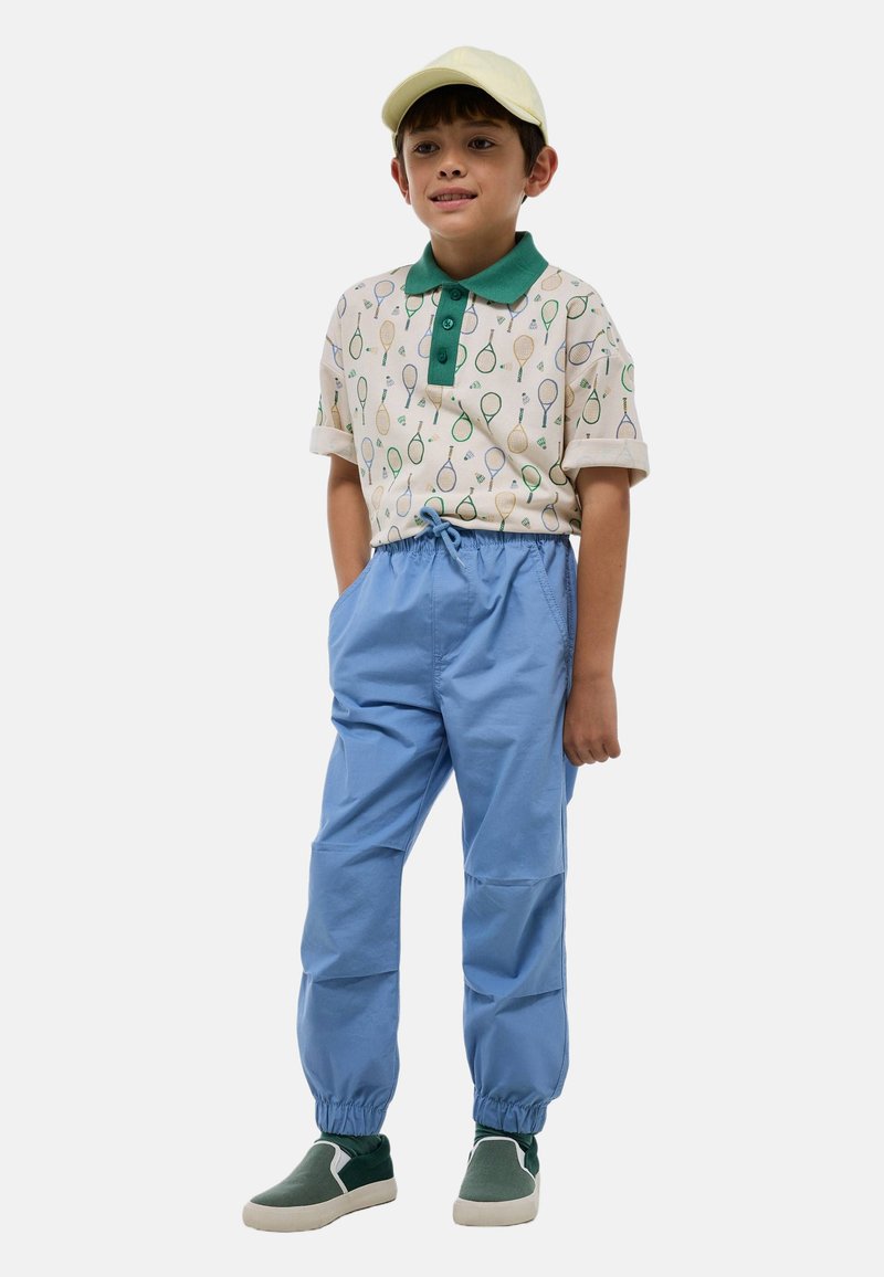 Polo à manches courtes avec col vert et motif de raquette de tennis, associé à un pantalon bleu à taille élastique et des chaussures vertes à enfiler. Casquette jaune.