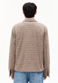 Bruin geruite jas met een zachte textuur, voorzien van een kraag en zijsplitten. Gecombineerd met blauwe denim, wat een relaxte pasvorm laat zien.