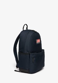 Sac à dos bleu marine avec poignée supérieure, bretelles rembourrées, poche avant zippée, et un patch arborant un drapeau américain et le texte "Superdry".