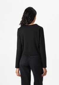 Schwarzes Langarmoberteil mit rundem Ausschnitt, strukturiertem Stoff und cropped Design. Getragen mit dunklen Jeans, was einen einfachen und modernen Look zeigt.