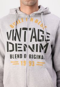 Szürke zipzáras kapucnis felső, elülső zsebbel, húzózsinórokkal, és merész fekete és sárga felirattal: "Built in Blue Vintage Denim Blend Original 1993."