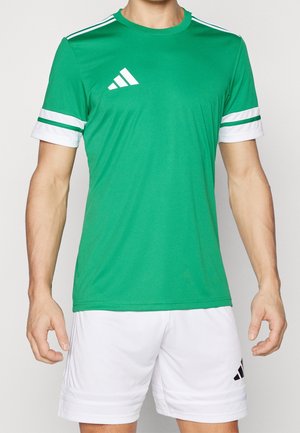 Homme portant un t-shirt de sport vert Adidas avec des bandes blanches sur les manches et un short Adidas blanc sur un fond uni.
