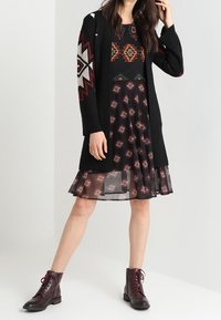 Cardigan nero a maglia con motivi geometrici sulle maniche, indossato sopra un vestito nero con motivi circolari rossi e verdi. Stivali marroni con allacciatura.