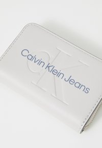 Pilkas sintetinės odos pinigin�ė su iškilniu "CK" logotipu ir mėlynu tekstu "Calvin Klein Jeans", pasižyminti lygiu paviršiumi ir siuvinėtomis kraštinėmis.