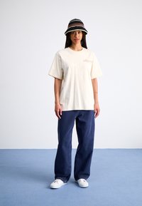 Kremfarget bomull T-skjorte med korte ermer og subtilt logo, parret med mørkeblå vide jeans og en svartstripete bøttehatt.