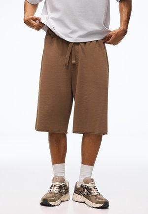 Mężczyzna noszący brązowe krótkie spodenki sięgające kolan z troczkiem, białe skarpetki, beżowe trampki oraz białą koszulę lekko podniesioną, aby pokazać pas.