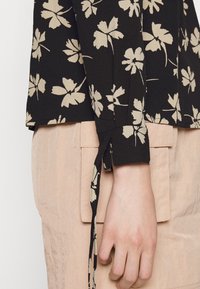 Blusa nera con motivo floreale beige, con dettaglio a fiocco al polso, abbinata a un capo di abbigliamento rosa chiaro e testurizzato.
