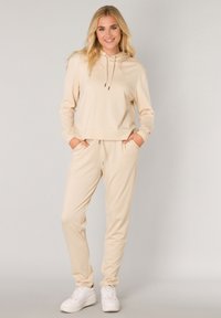 Beige hoodie met voorzakken en trekkoorden, gecombineerd met bijpassende taps toelopende broek en witte sneakers. Zachte, gladde stof met een minimaal ontwerp.