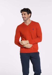 Pull à col en V, à manches longues, en tricot orange vif, avec un petit logo bleu sur la poitrine gauche. Porté avec un pantalon de couleur marine.