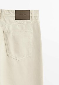 Beige bomullsbyxor med en bakficka, sydda detaljer och en mörkbrun läderlogga vid midjan.