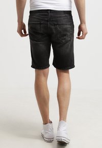 Persona vistiendo pantalones cortos de mezclilla negros, zapatillas Converse altas blancas y una camisa blanca, de pie de espaldas sobre un fondo claro.