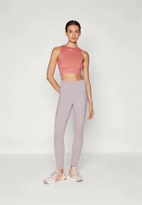 Femme se tenant avec une main derrière le dos, portant un top de sport sans manches et court, un legging taille haute avec poches et des chaussures de sport.