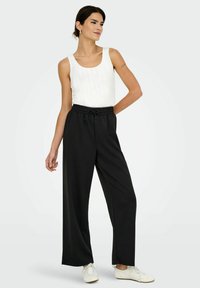 Schwarze weit geschnittene Hose mit einem elastischen Bund und Kordelzug, kombiniert mit einem weißen gerippten Tanktop. Die Schuhe sind weiß mit Schnürsenkeln.