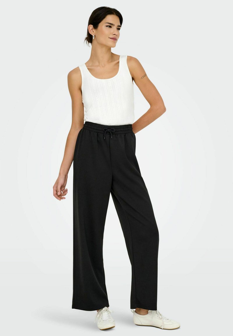 Schwarze weit geschnittene Hose mit einem elastischen Bund und Kordelzug, kombiniert mit einem weißen gerippten Tanktop. Die Schuhe sind weiß mit Schnürsenkeln.