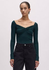 Dunkelgrüner Langarm-Bodysuit mit herzförmigem Ausschnitt und Twist-Detail an der Vorderseite, kombiniert mit hochgeschnittenen schwarzen Jeans.