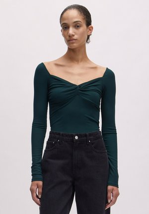 Donkergroen langhanskleding met een sweetheart neckline en een twistdetail aan de voorkant, gecombineerd met hoge zwarte jeans.