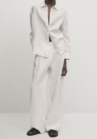 Chemise blanche en lin à manches longues et pantalon à jambes larges, présentant une coupe décontractée et une texture subtile. Sandales plates noires complètent la tenue.