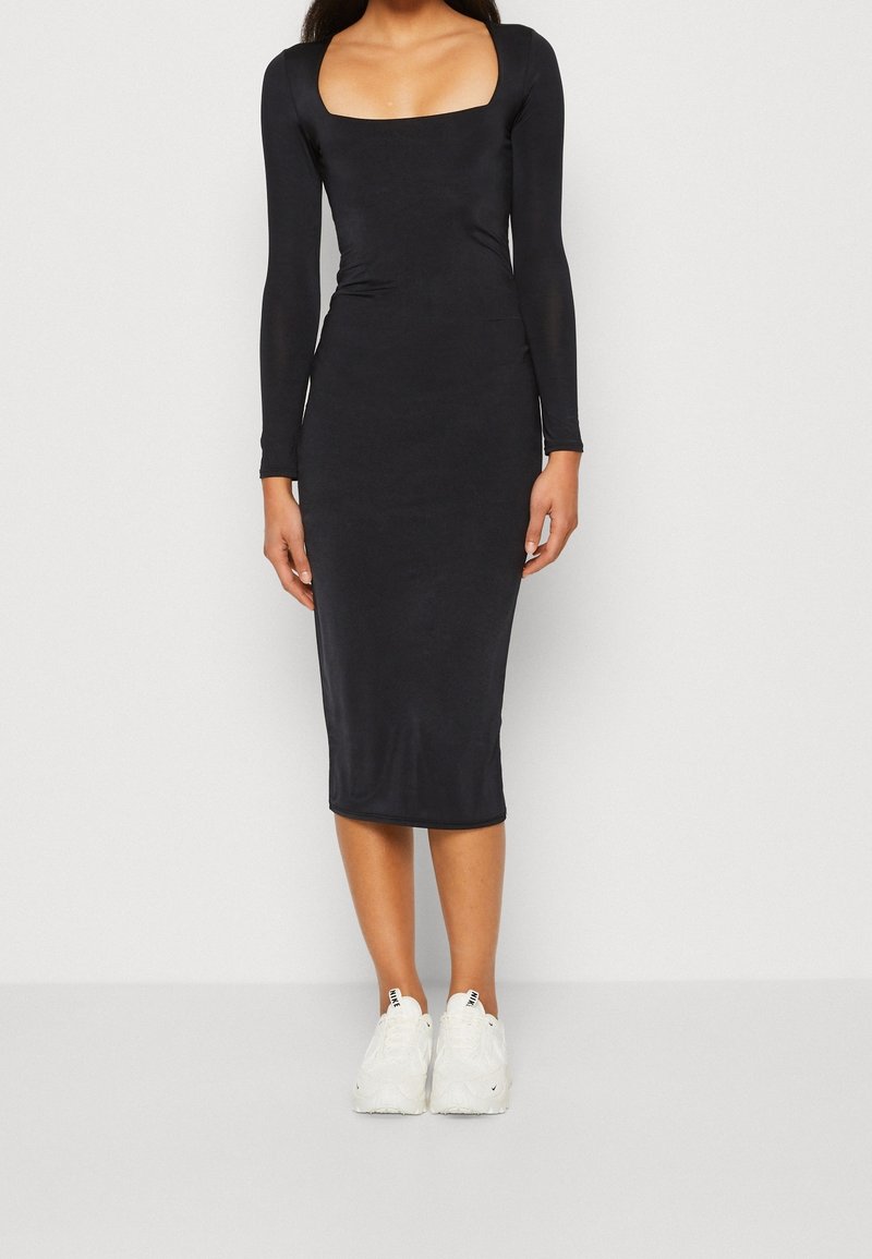 Femme portant une robe midi noire ajustée à manches longues avec un décolleté carré et des chaussures de sport blanches, debout contre un fond uni.