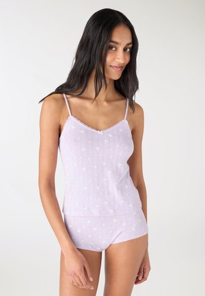 Giovane donna che indossa una canottiera floreale viola chiaro e pantaloncini abbinati, in piedi davanti a uno sfondo bianco, sorridendo dolcemente.