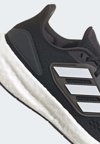 Czarne buty sportowe z teksturowaną cholewką, białą podeszwą amortyzującą i charakterystycznymi czarno-białymi paskami po bokach. Okrągły nos.