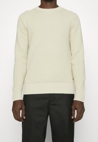 Cremefarbener Strickpullover mit strukturiertem Muster, Rundhalsausschnitt und gerippten Bündchen. Kombiniert mit dunklen Hosen, die eine kontrastierende Farbpalette zeigen.