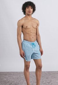 Mladý muž s kučeravými vlasmi stojaci bosý, oblečený v modrých plavkách so vzorom a tyrkysovým sťahovacím pásom na pozadí bez vzoru.