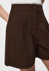 Shorts marron foncé taille haute, confectionnés sur mesure avec plis devant, fermeture par bouton et poches latérales, portés avec un haut blanc.