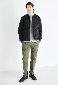 Chaqueta negra acolchada con botones a presión, sudadera gris debajo, pantalones verdes y zapatillas negras, todo sobre un fondo blanco liso.