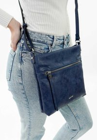 Borsa a tracolla blu navy in pelle testurizzata, con una tasca frontale con zip e una tracolla regolabile. Indossata insieme a jeans azzurri.