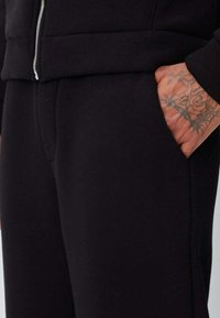 Joggers en tissu noir doux avec une coupe ajustée. Caractéristiques : poches latérales et taille côtelée, illustrés par une main reposant sur une poche.