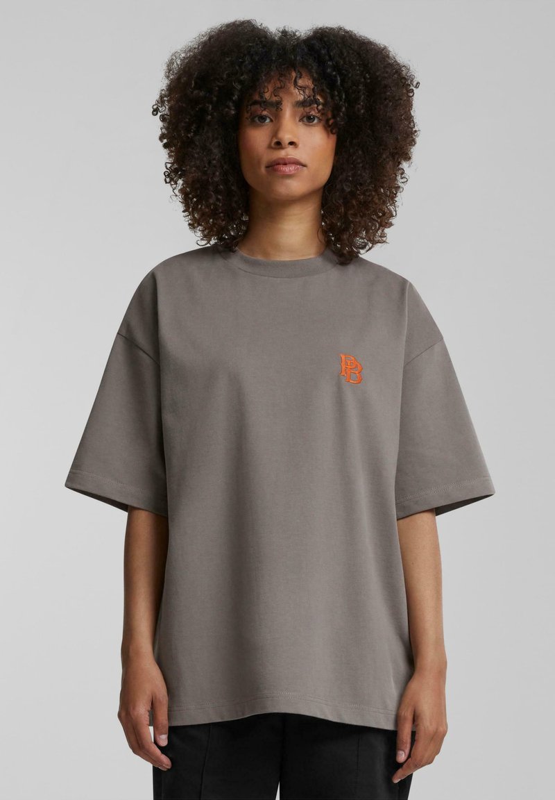 Camiseta gris de gran tamaño hecha de algodón, con mangas cortas, cuello redondo y un pequeño logotipo naranja en el lado izquierdo del pecho.
