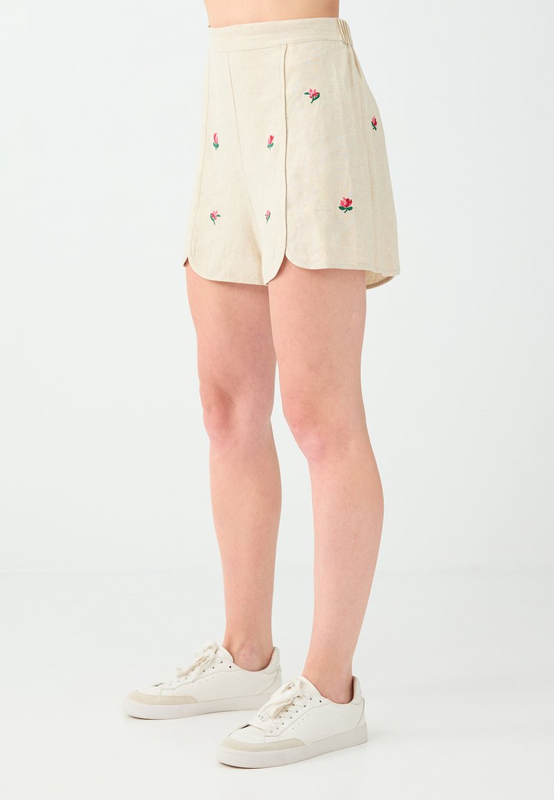 Short beige avec une petite broderie de fleur rose, porté avec des baskets blanches basses sur une personne debout devant un fond uni.